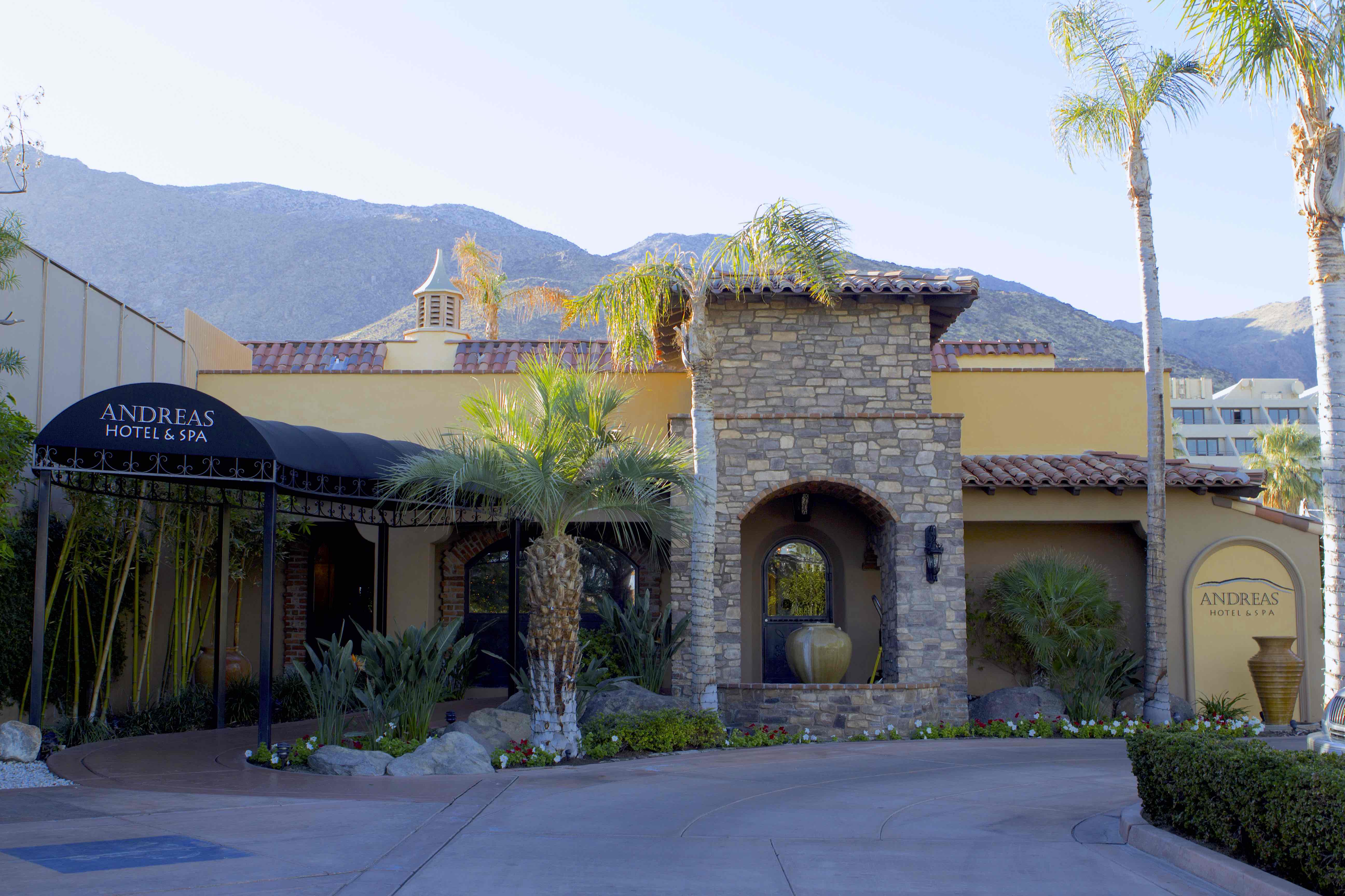 A stone and yellow stucco building with a tile roof, palm trees, and a mountain view.
